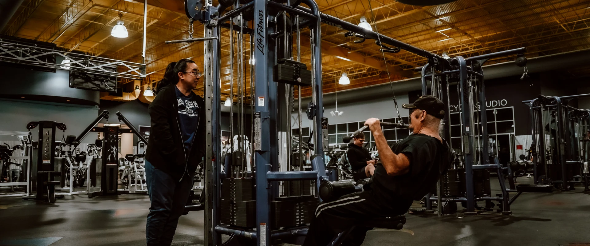 A man using a cable weight machine for seated lat pulldowns at Gold’s Gym, Walker Springs, Tennessee. Another individual stands nearby observing. The gym features industrial-style lighting, blue fitness equipment, and a spacious layout with additional machines and mirrors in the background. The bright yellow ceiling adds warmth to the modern gym environment.