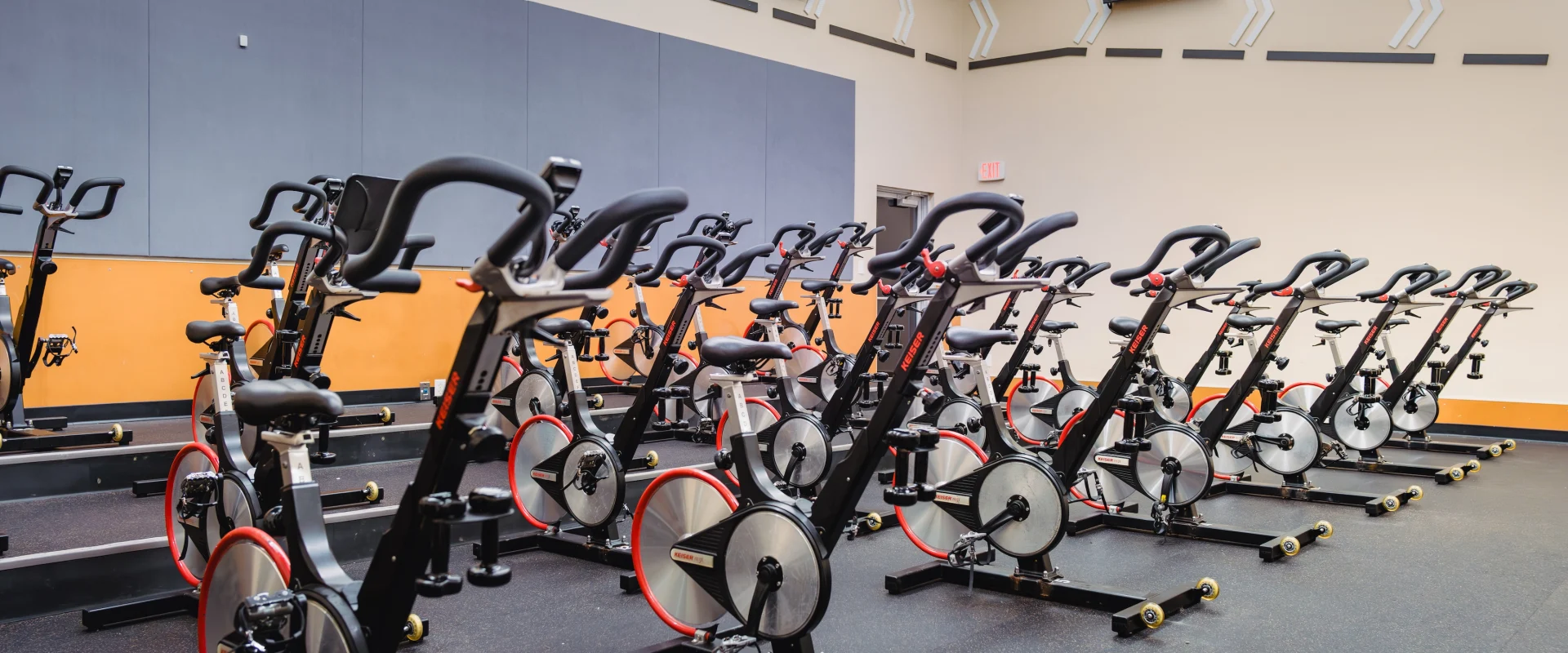 A bright, well-lit gym room with multiple stationary bikes neatly arranged in rows on a black rubber floor, creating a clean and organized atmosphere.