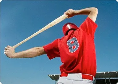 A person in a red baseball jersey is swinging a baseball bat against a clear blue sky background.