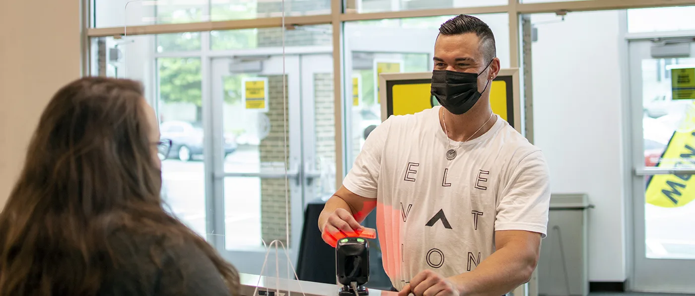 A man wearing a white t-shirt with text and a face mask is standing in front of a glass window, interacting with a woman in the background.