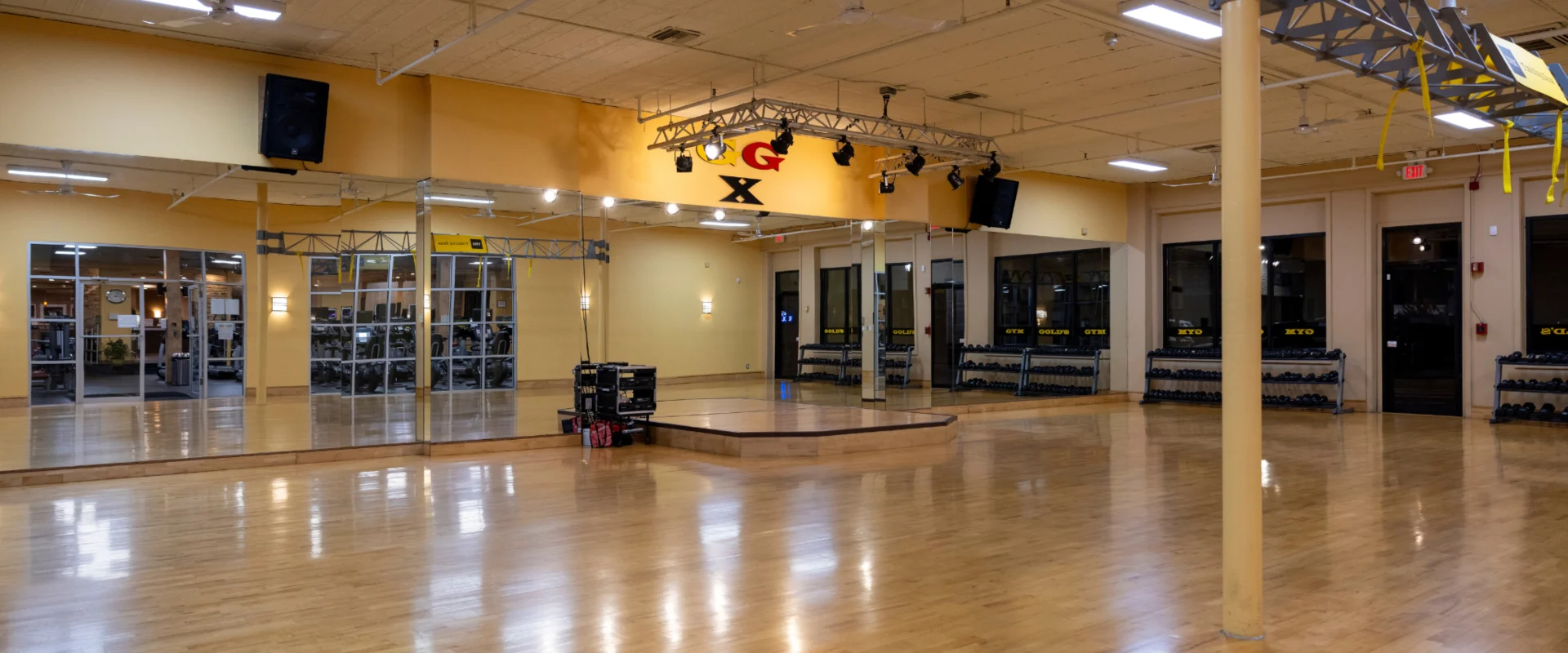 Group exercise studio at Gold’s Gym in East Northport, New York, featuring a spacious hardwood floor, mirrored walls, and a raised instructor stage. The room includes TRX suspension trainers, stacked dumbbells along the perimeter, and ceiling-mounted lighting for an energetic class atmosphere. Large windows provide natural light, with views of the main gym area visible through glass panels.