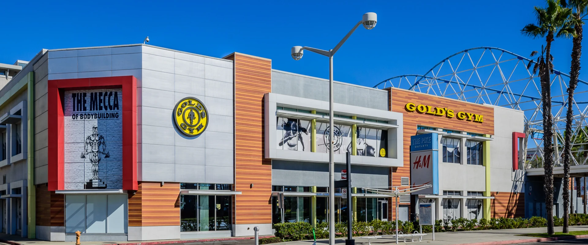 Exterior of Gold’s Gym Long Beach, showcasing a modern multi-story design with large windows and bold signage. The facade features wood paneling, sleek metal accents, and a prominent mural labeling it as "The Mecca of Bodybuilding." A covered walkway and nearby retail stores integrate the gym into a bustling shopping center. Palm trees and a bright blue sky enhance the vibrant, coastal setting.