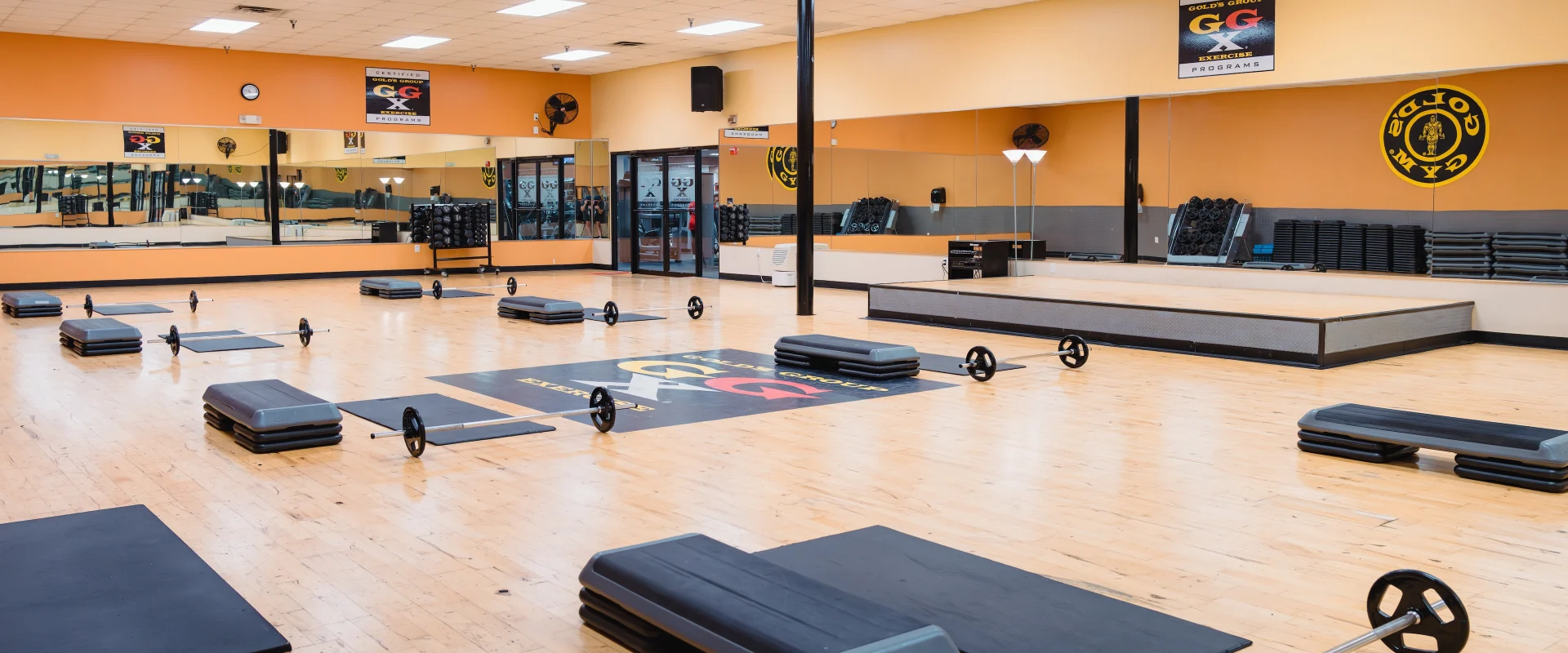 Group exercise studio at Gold’s Gym in Austin (Southeast), set up with barbells, aerobic steps, and mats arranged neatly on a polished wooden floor. A mirrored wall reflects the spacious room, with stacked equipment along the perimeter for fitness classes.