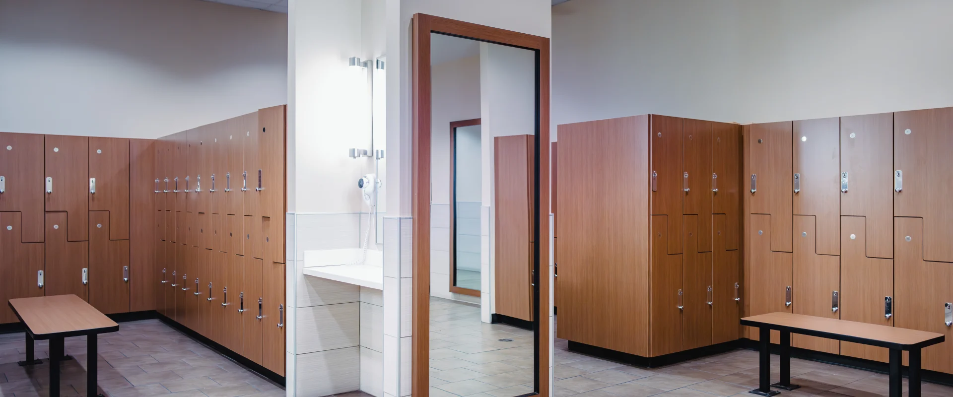 A clean locker room at Gold's Gym with wooden lockers, tiled flooring, benches, a full-length mirror, and a vanity area equipped with a hairdryer.