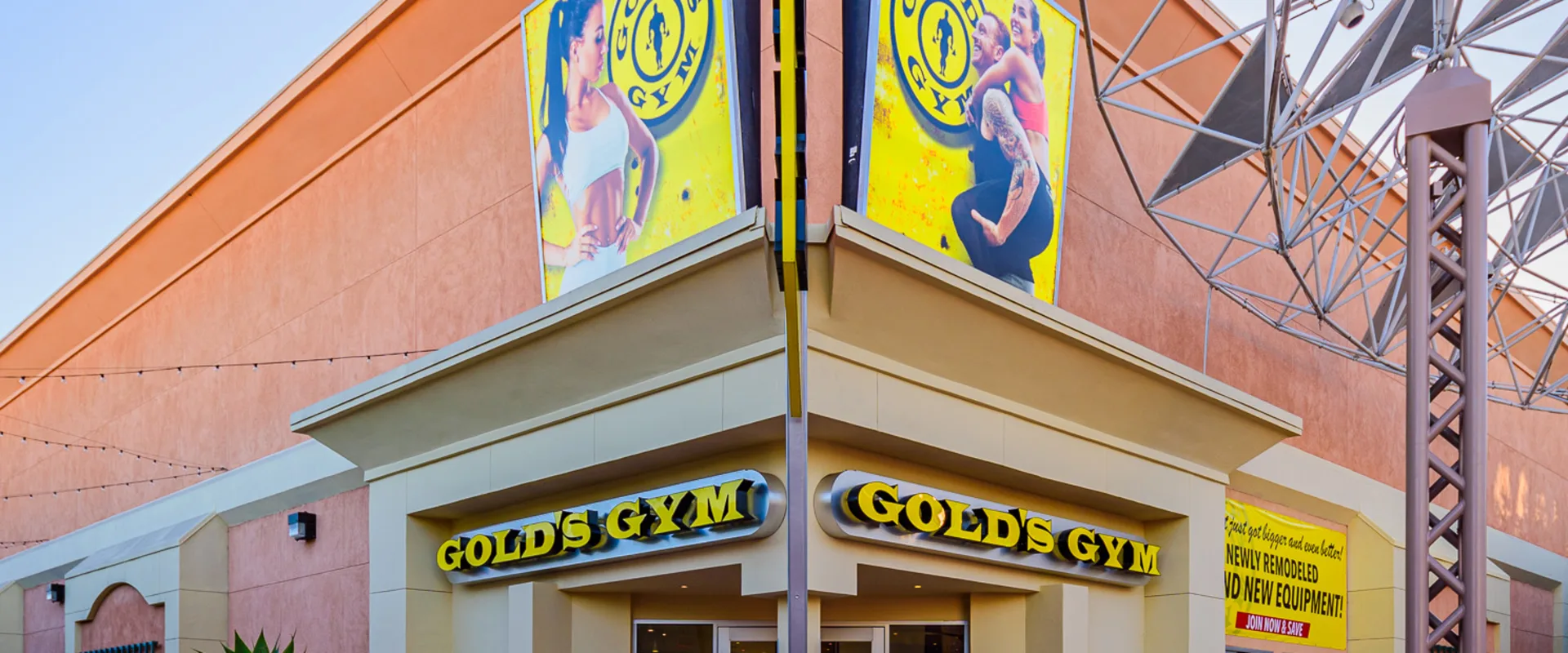 Exterior view of Gold’s Gym Thousand Oaks, showing the entrance with double glass doors framed by beige walls. Above the doors, two illuminated Gold’s Gym signs are mounted on the building’s facade. Large promotional banners with gym branding and fitness imagery are displayed on the upper walls. A potted plant and bicycles are positioned near the entrance. The structure features an open-air metal framework extending from the roof. String lights are visible along the side of the building.