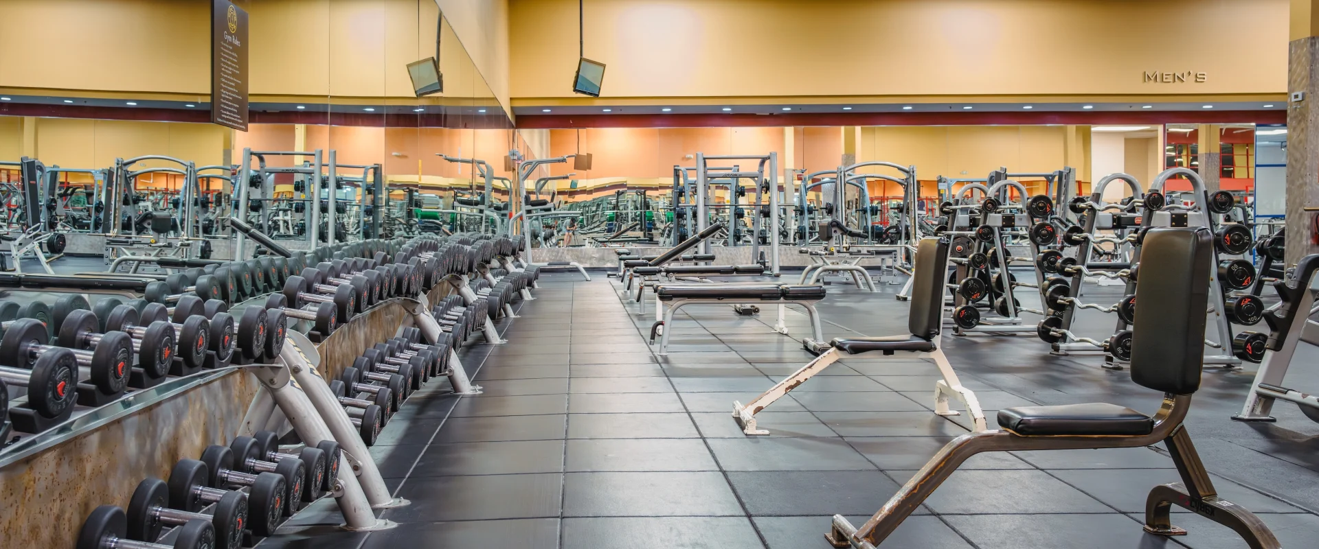 A spacious gym with rows of dumbbells, benches, and various workout machines. The room has a mirror wall, dark flooring, and a bright, inviting atmosphere.