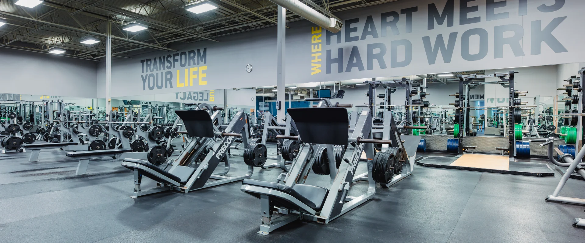 Plate-loaded strength training area at Gold's Gym Waco featuring leg press machines, squat racks, and benches with ample weight plates nearby. The space includes mirrored walls, motivational text on the walls, and rubber flooring for a durable workout environment. Ceiling lighting provides bright, even illumination.