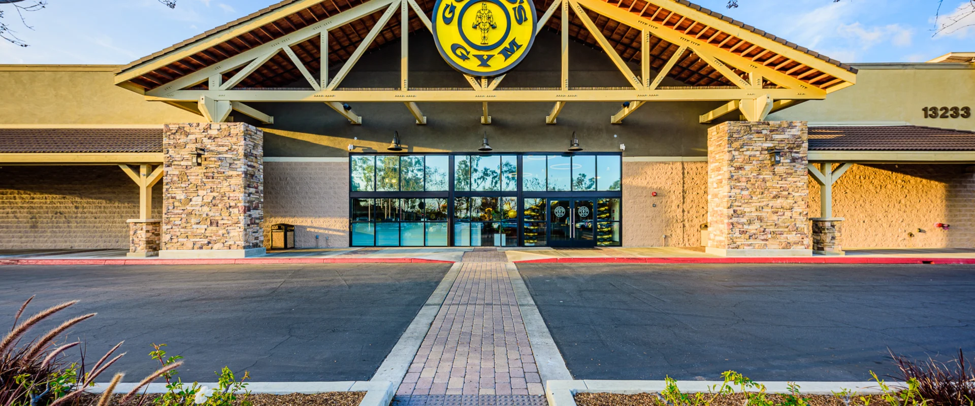 Exterior of Gold’s Gym Cerritos, featuring a grand entrance with a high-pitched roof supported by exposed wooden beams. A large Gold’s Gym logo is displayed above the glass doors, reflecting the surrounding trees and sky. The stone-clad columns and warm-toned facade create a welcoming aesthetic. A paved walkway leads to the entrance, lined with landscaped greenery.
