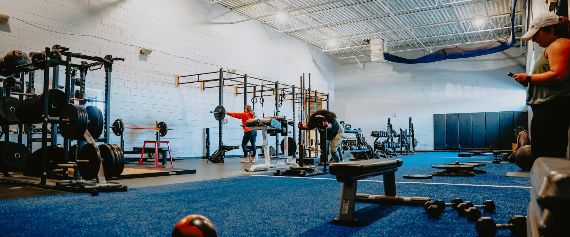 Functional training area at Gold’s Gym Tennessee, Lee Highway, featuring a wide-open space with vibrant blue turf flooring. The area includes racks of weights, barbells, and functional equipment like gymnastic rings and medicine balls. A variety of functional stations and benches are arranged across the space, with members engaged in exercises. The white walls and high ceilings with exposed beams and bright overhead lighting create an airy, focused workout environment.