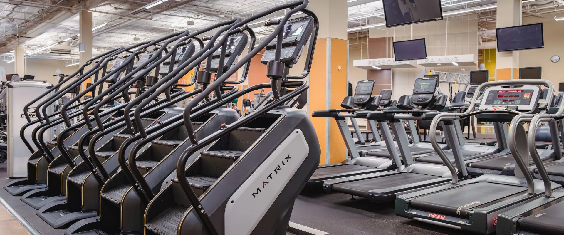 Cardio area at Gold’s Gym in Austin (Westlake), featuring a row of Matrix stair climbers alongside treadmills equipped with digital screens. Overhead TVs are mounted for entertainment during workouts, while the open ceiling design and bright lighting create an airy, spacious environment. The area includes rubber flooring for added safety and support during cardio sessions.