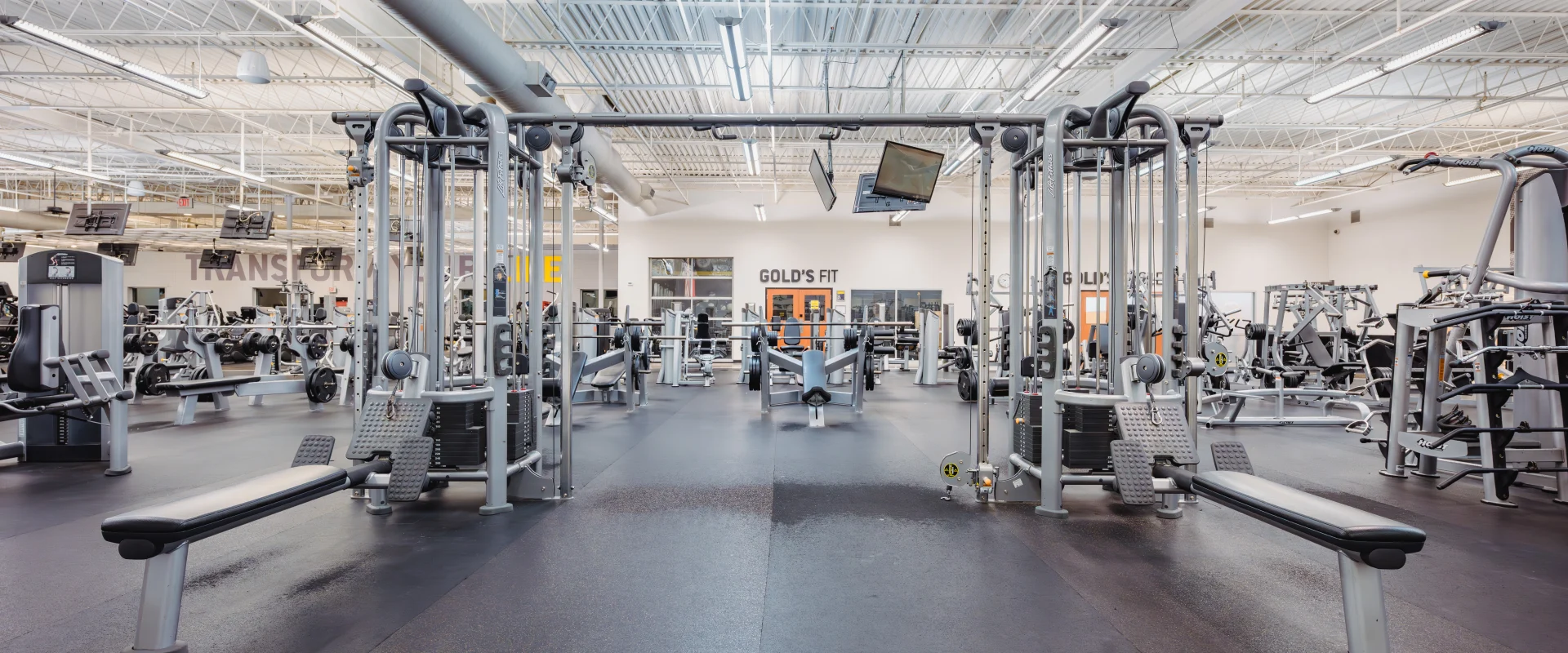 Spacious gym area featuring machine weight equipment arranged in rows on black rubber flooring. Centralized cable machines are flanked by benches and adjustable seats, while additional strength-training machines are visible in the background. The high ceiling with exposed ductwork enhances the open atmosphere, with overhead fluorescent lighting providing even illumination. A "Gold's Fit" sign and windows are visible in the rear wall, adding branding and separation to the space. Monitors hang from the ceiling for entertainment.