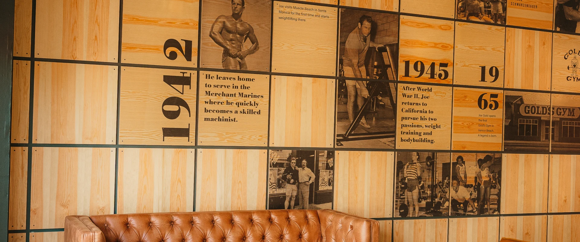Lounge area at Gold's Gym Hendersonville featuring a tufted leather couch, two orange chairs, a round wooden coffee table, and a wall decorated with historic Gold's Gym milestones.