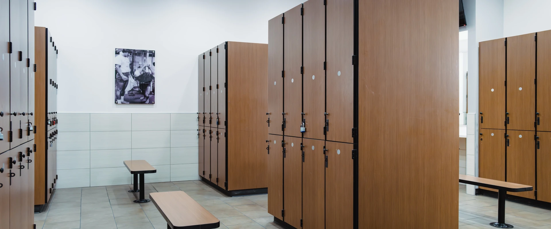 Locker room at Gold's Gym in Texas (Pflugerville), featuring rows of wooden lockers with black trim, secure locks, and metal benches for seating. The space has tiled floors, white tiled walls, and bright overhead lighting for a clean, organized look.