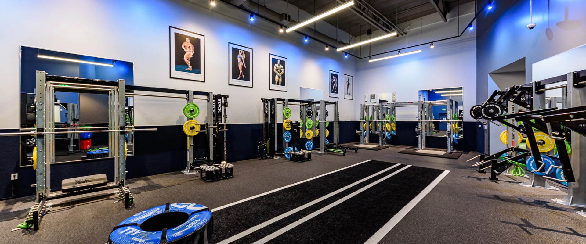 An area of the gym with black turf and weighted tires at the end on the left. Outside of the turf are various weightlifting racks with various weight.