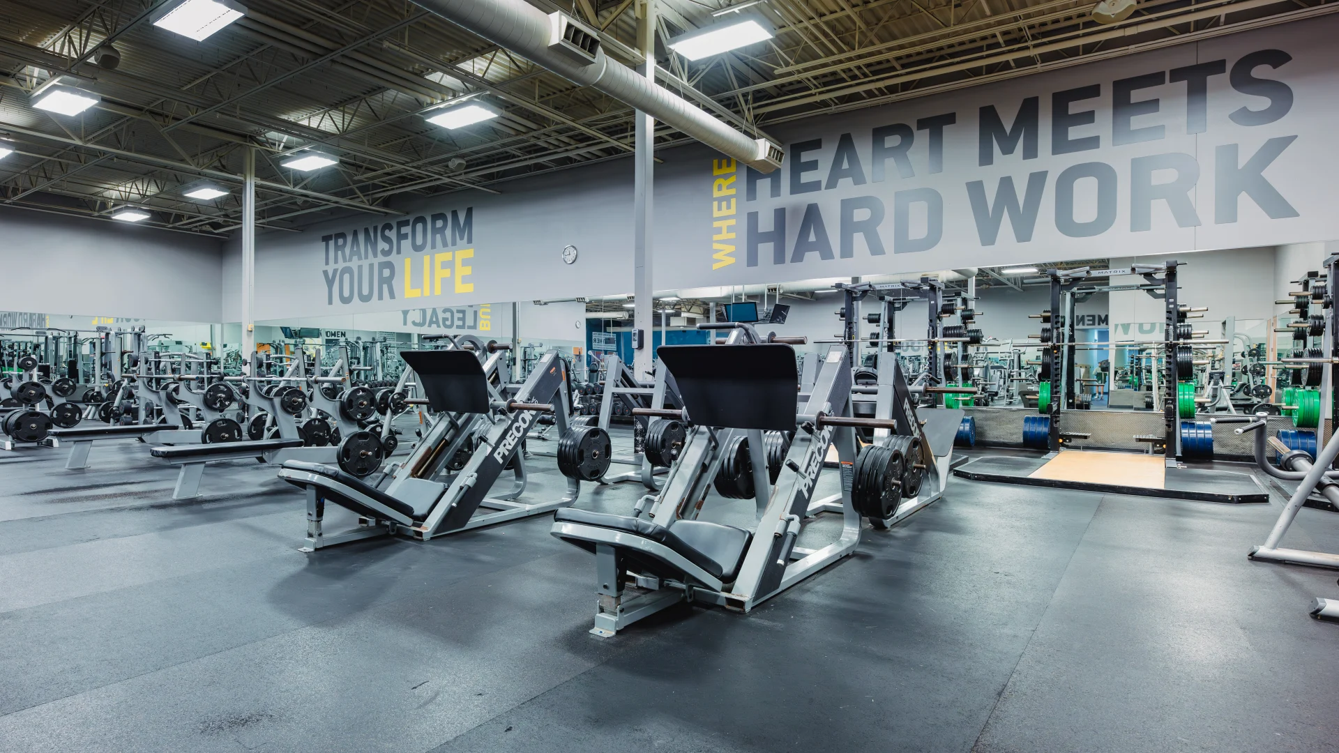 Plate-loaded strength training area at Gold's Gym Waco featuring leg press machines, squat racks, and benches with ample weight plates nearby. The space includes mirrored walls, motivational text on the walls, and rubber flooring for a durable workout environment. Ceiling lighting provides bright, even illumination.