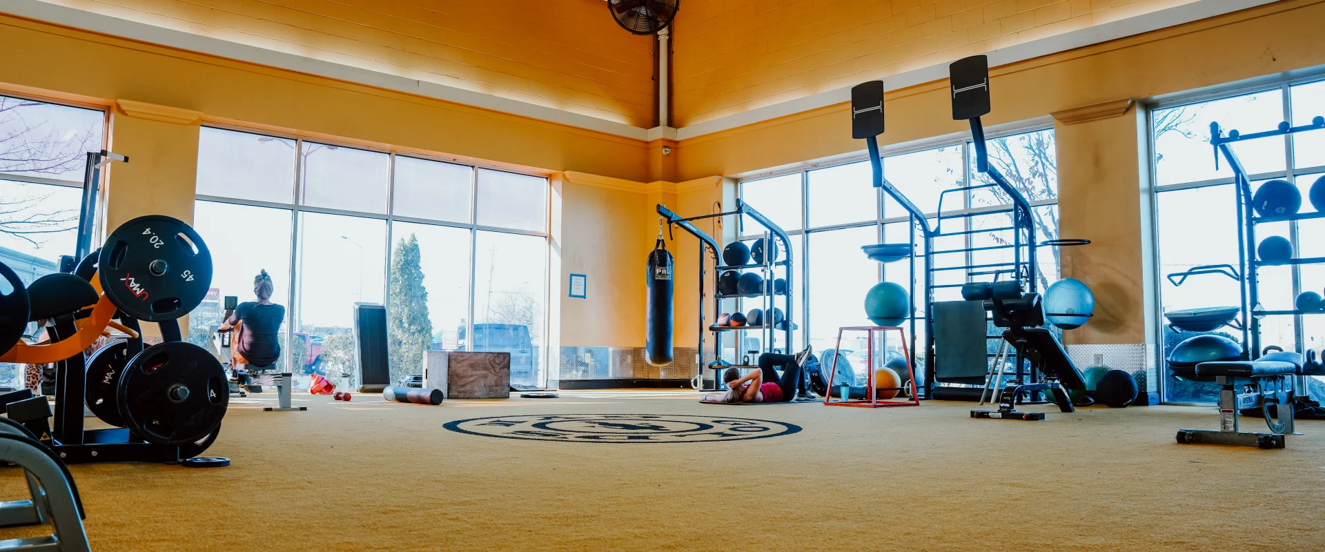 Functional training area at Gold's Gym Murfreesboro, Tennessee, featuring a yellow carpet, bright yellow walls, and a ceiling-mounted industrial fan. The space includes a punching bag, weight plates, medicine balls, kettlebells, a rowing machine, plyometric boxes, and a squat rack, with large windows providing natural light.