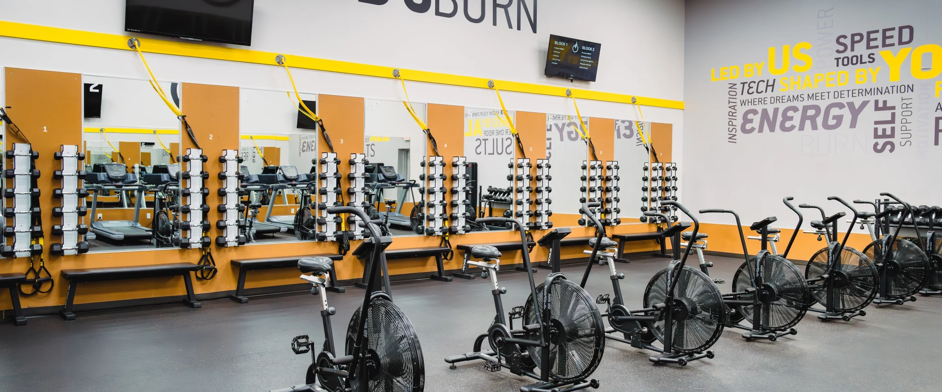 Gold's Burn studio at Gold’s Gym in Texas (N. Round Rock), featuring a row of air bikes positioned in front of mirrored walls. Dumbbell racks with neatly organized weights line the mirrored wall, accompanied by TRX suspension straps anchored above. Flat benches are set below the mirrors for strength training exercises. The space includes motivational wall graphics and ceiling-mounted TVs for workout guidance. The flooring is rubberized, providing durability and shock absorption.