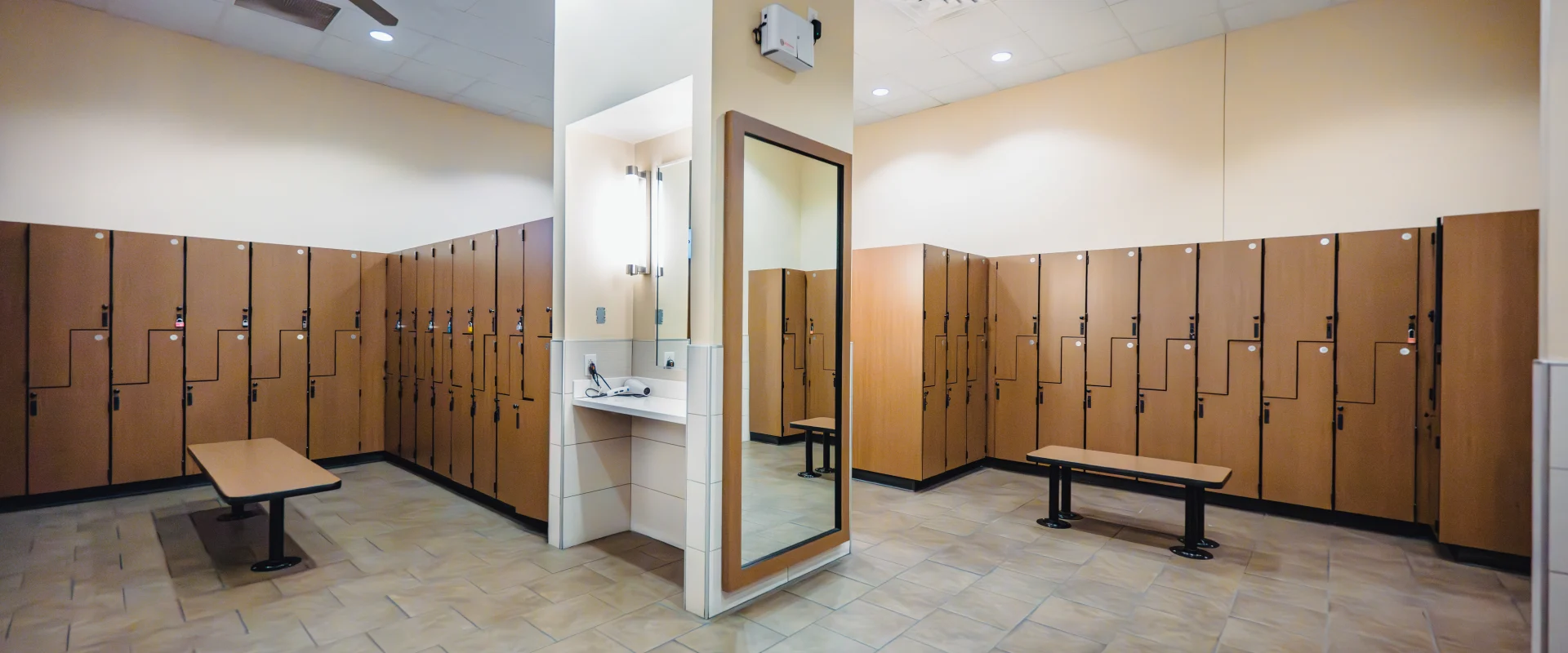 Spacious locker room at Gold's Gym featuring wooden lockers, tiled floors, benches, and a mirror with a vanity area and bright overhead lighting.