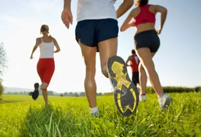 Three people in athletic clothing are running through a grassy field with a scenic landscape in the background.