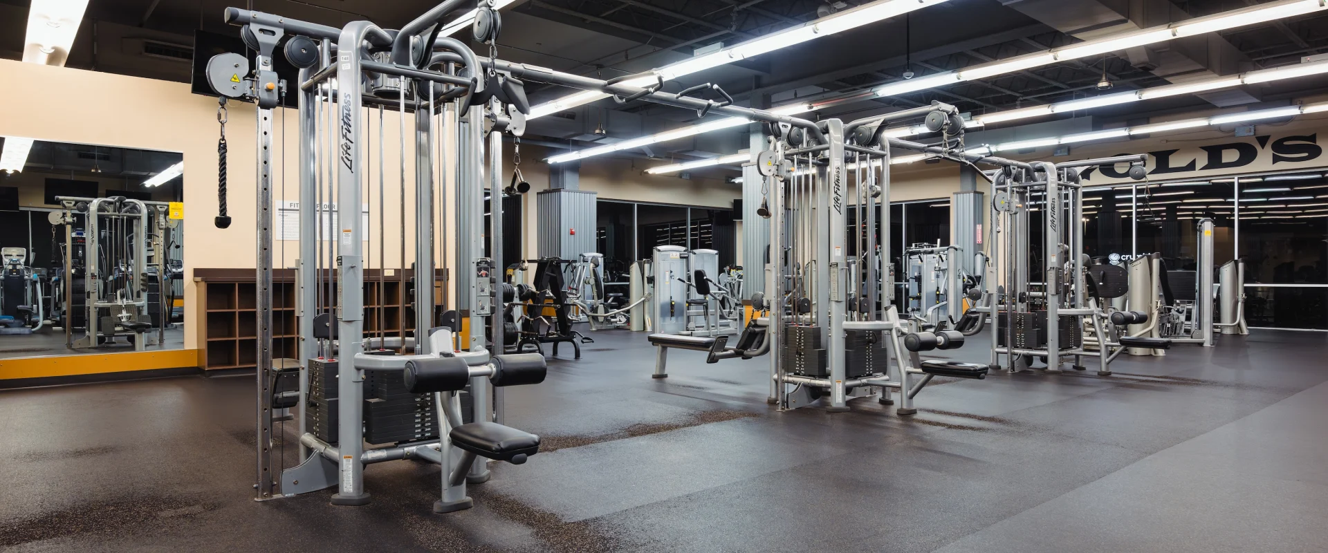 A spacious gym with various exercise machines, well-lit with overhead lights, mirrors on the walls, and a clean, modern appearance. The space is empty.