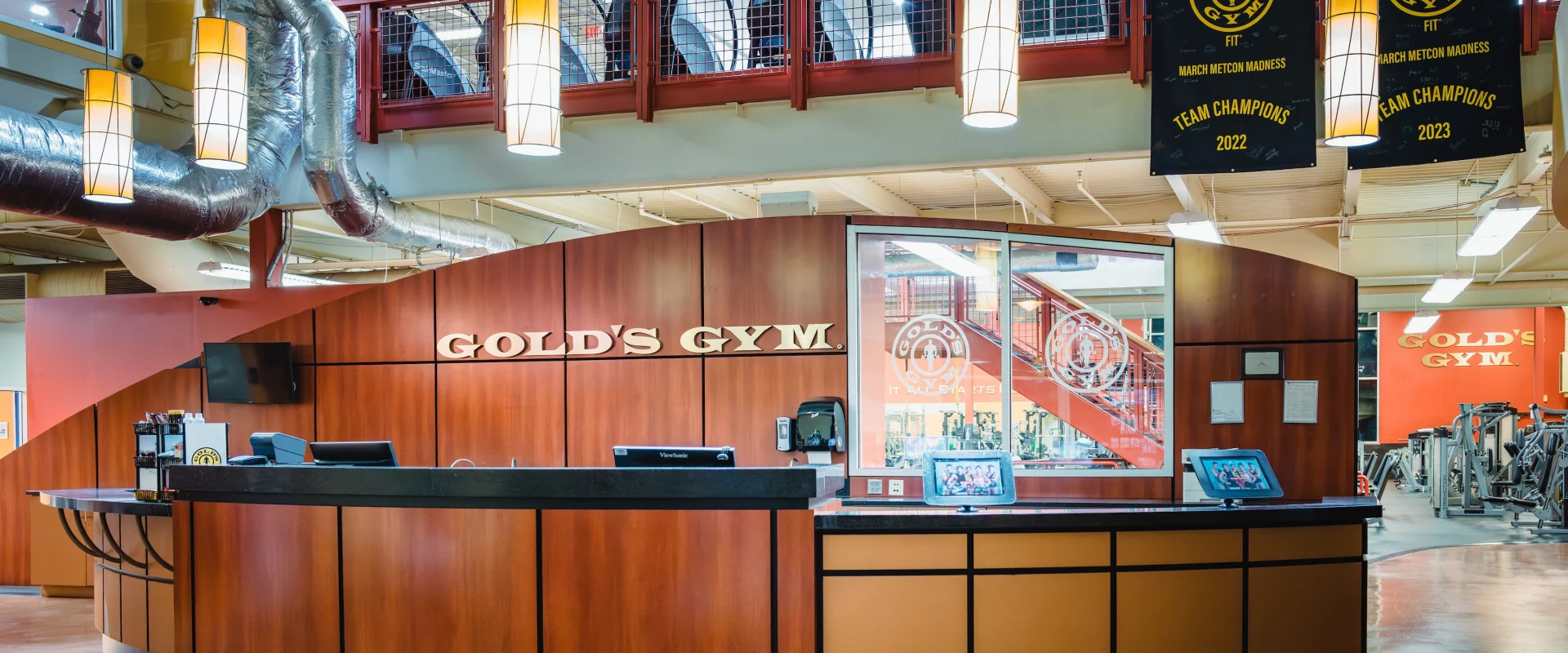 Reception area of Gold's Gym Fiesta Trails with a large wooden desk. Above, there's a balcony with exercise equipment and two championship banners.