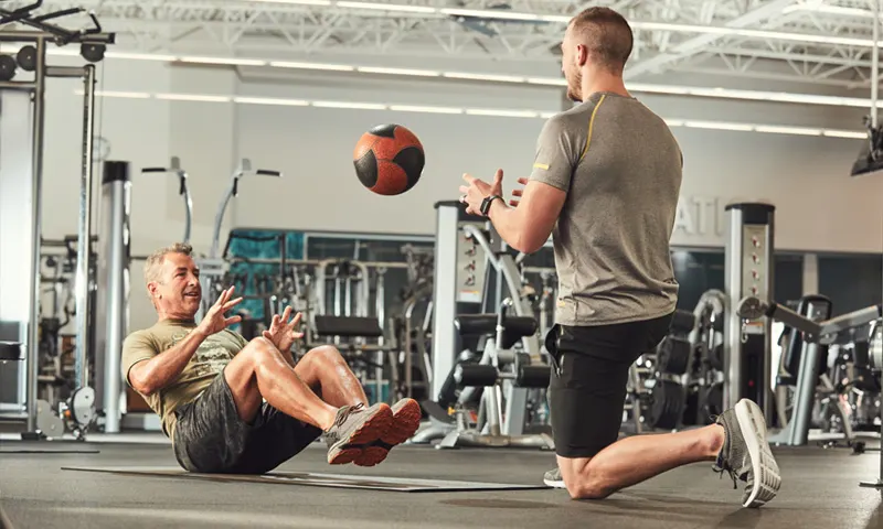 Two individuals are engaged in a workout routine in a well-equipped gym setting, with various fitness equipment and machines visible in the background.