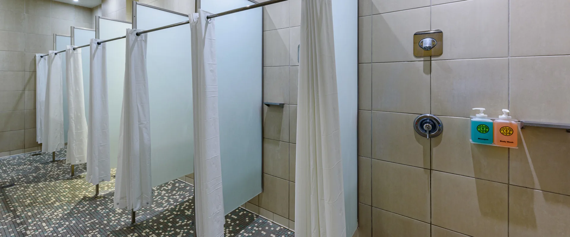 Shower area in the locker rooms at Gold’s Gym Thousand Oaks, featuring multiple private stalls with frosted dividers and white curtains for privacy. Beige tiled walls and mosaic flooring create a clean, modern look. Wall-mounted dispensers provide shampoo and body wash for convenience.