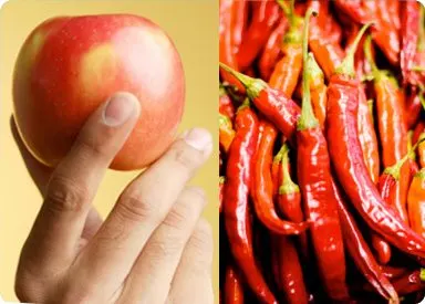 A hand holding a red apple, and a pile of vibrant red chili peppers in the background.