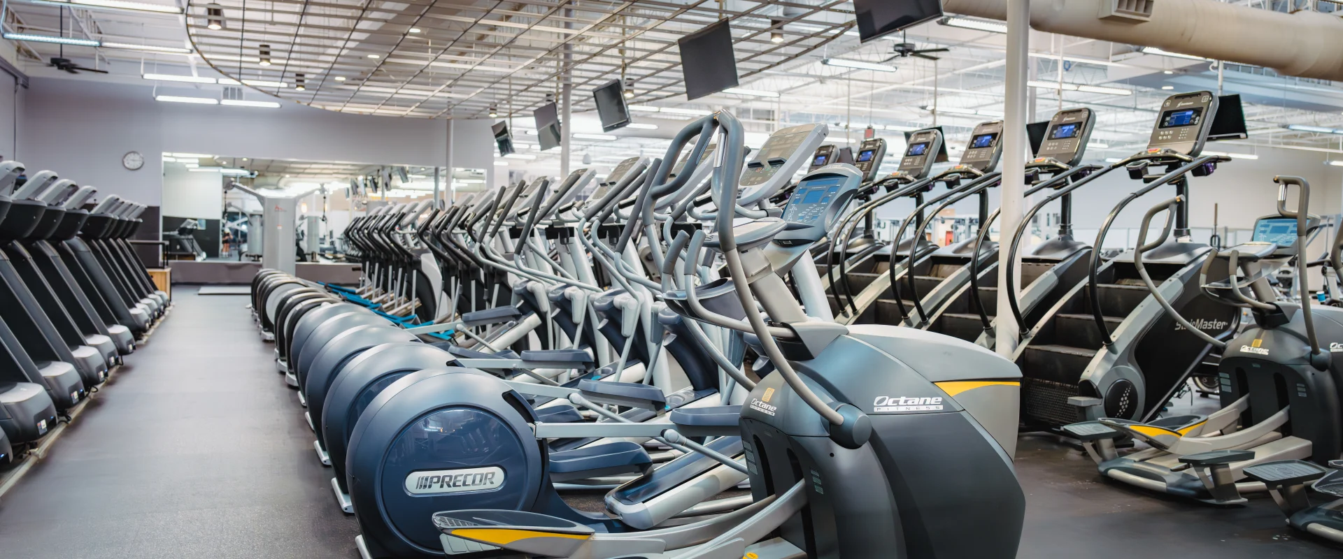 Cardio area at Gold's Gym in Austin (Hesters Crossing) featuring rows of elliptical machines and treadmills aligned on a sleek black floor. The space is brightly lit with overhead industrial-style lighting and features mirrors along the back wall to enhance the open feel. Mounted TVs hang from the ceiling, offering entertainment for gym members during workouts.
