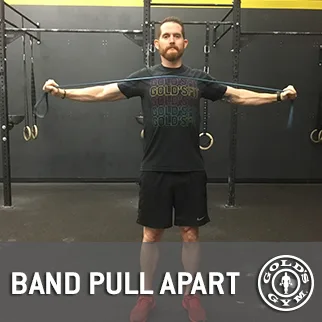 The image shows a man standing in a gym setting, wearing a black t-shirt with text, and performing a band pull-apart exercise with resistance bands.