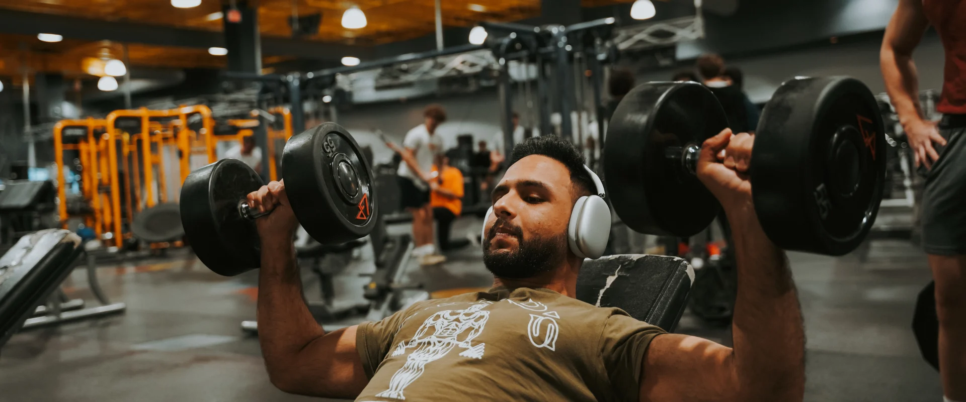 A man performing an incline dumbbell press in the free weights section at Gold’s Gym, Walker Springs, Tennessee. The gym features bright yellow strength-training racks in the background, with other members engaged in exercises. Overhead industrial-style lighting highlights the modern and energetic atmosphere of Gold’s Gym.