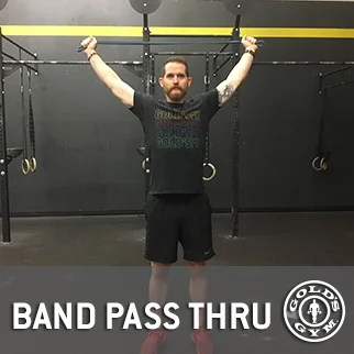 A man with a beard stands in a gym, raising his arms while wearing a black t-shirt with a logo, against a backdrop of exercise equipment and a "Band Pass Thru" sign.
