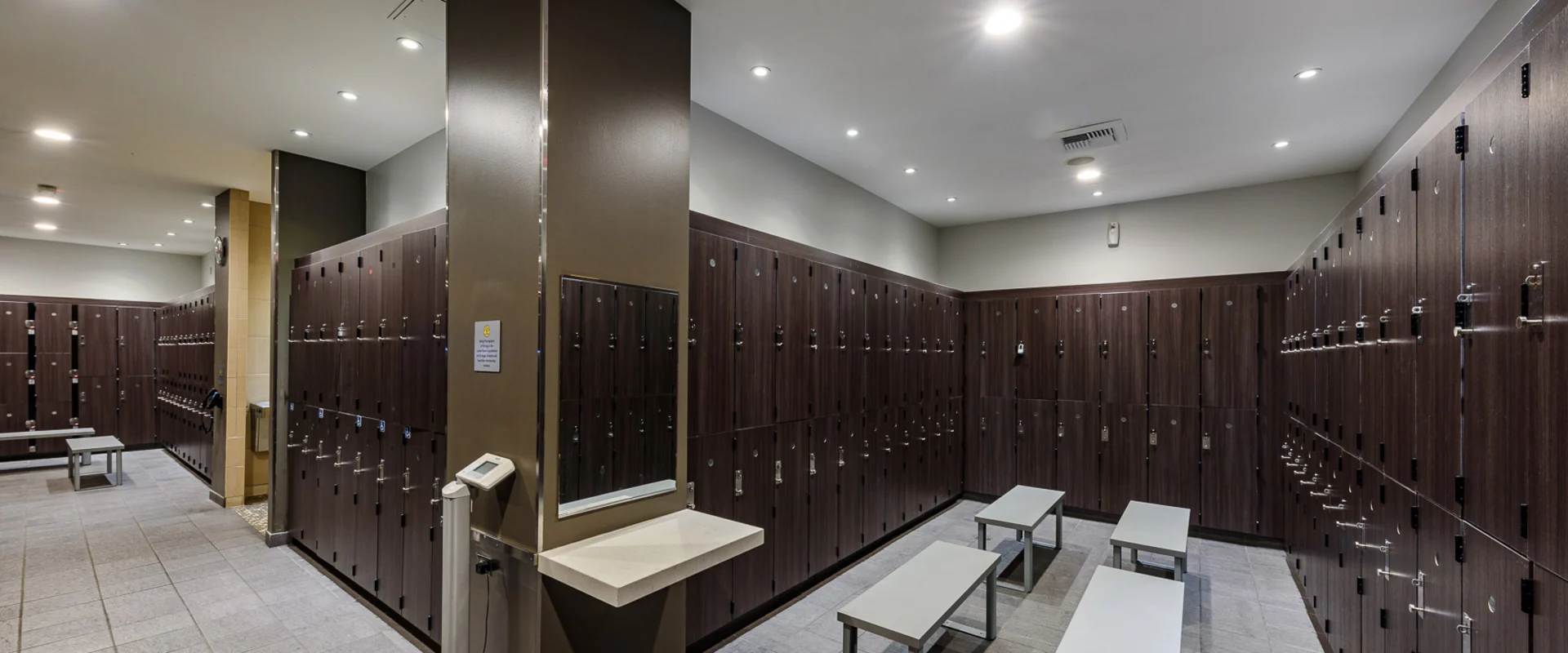 Gold’s Gym Thousand Oaks locker room featuring rows of dark wood lockers with built-in combination locks, light gray tile flooring, and sleek metal benches. A small vanity area with a mirror and countertop is positioned against a support column, adjacent to a digital scale. Recessed ceiling lights provide even illumination, enhancing the modern and functional design of the space.