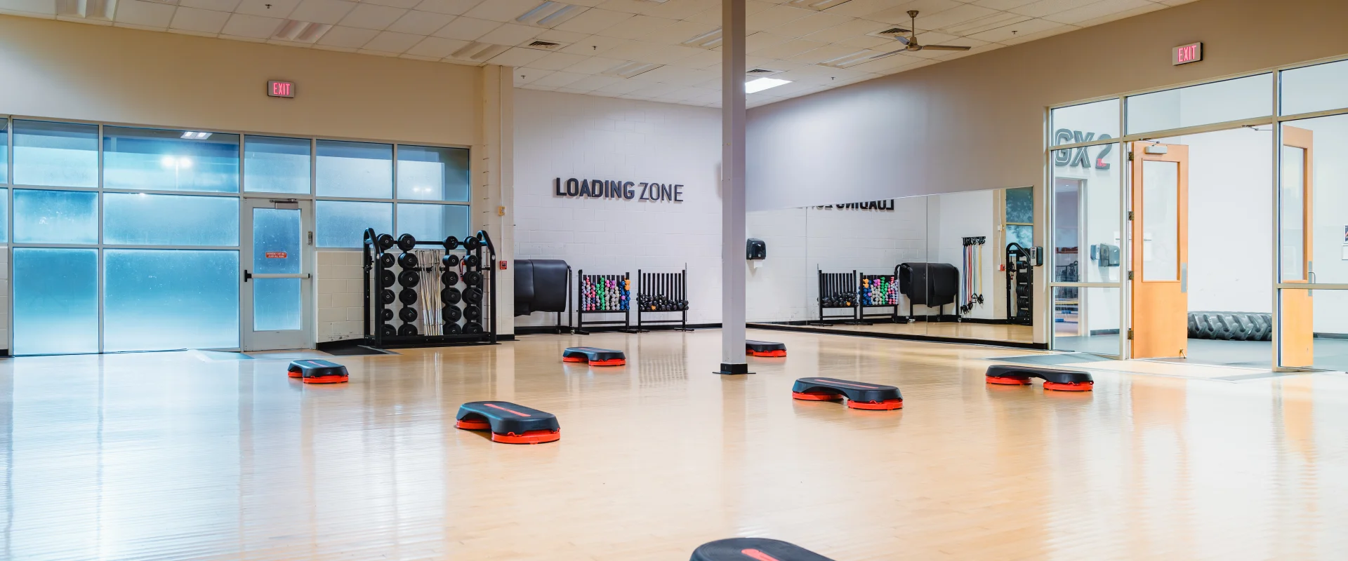 Spacious group exercise room at Gold's Gym Waco with a polished wood floor, step platforms evenly spaced, large mirrors along one wall, and storage racks holding weights and resistance bands. Frosted glass windows and double doors are visible.