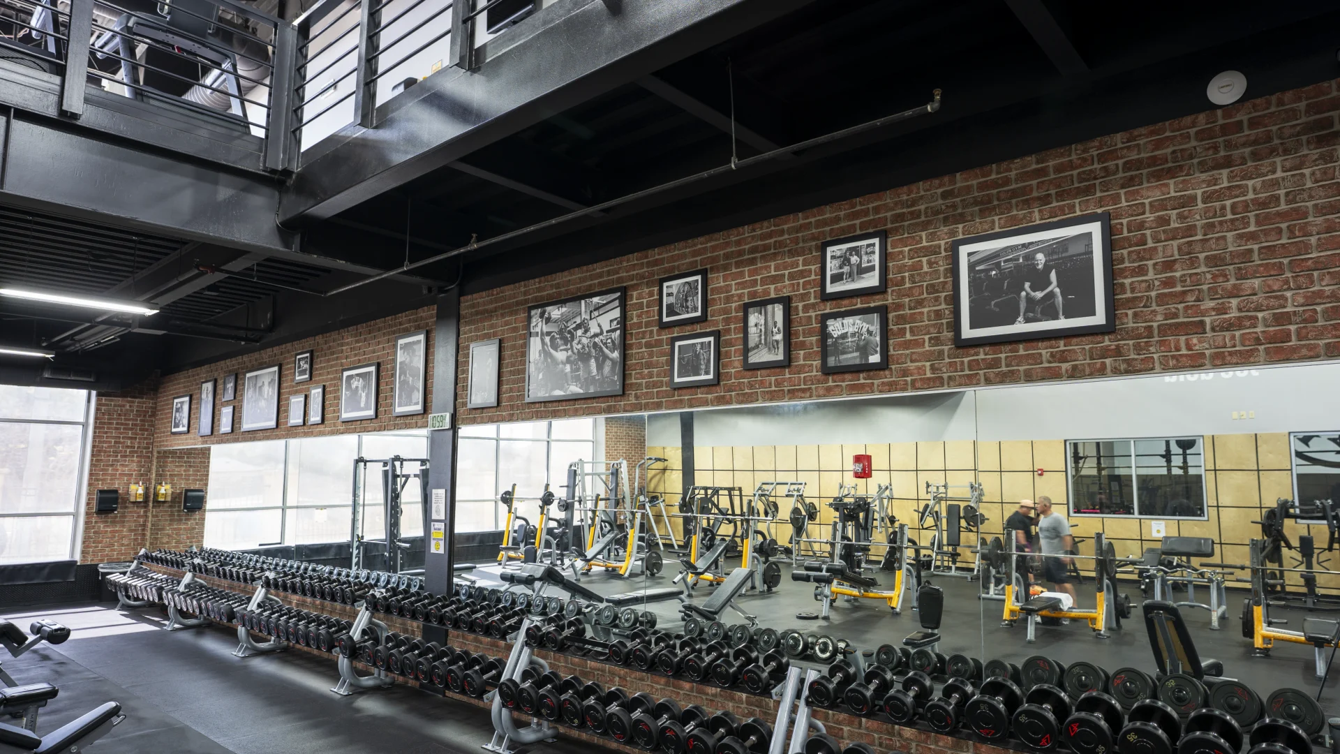 An interior view of a modern gym with a brick wall adorned with framed black-and-white photos of bodybuilders. A large mirror spans the length of the wall, reflecting rows of dumbbells on multi-tiered racks and various strength training machines. The space has an industrial design with black steel beams and a second-story mezzanine overlooking the main workout floor.