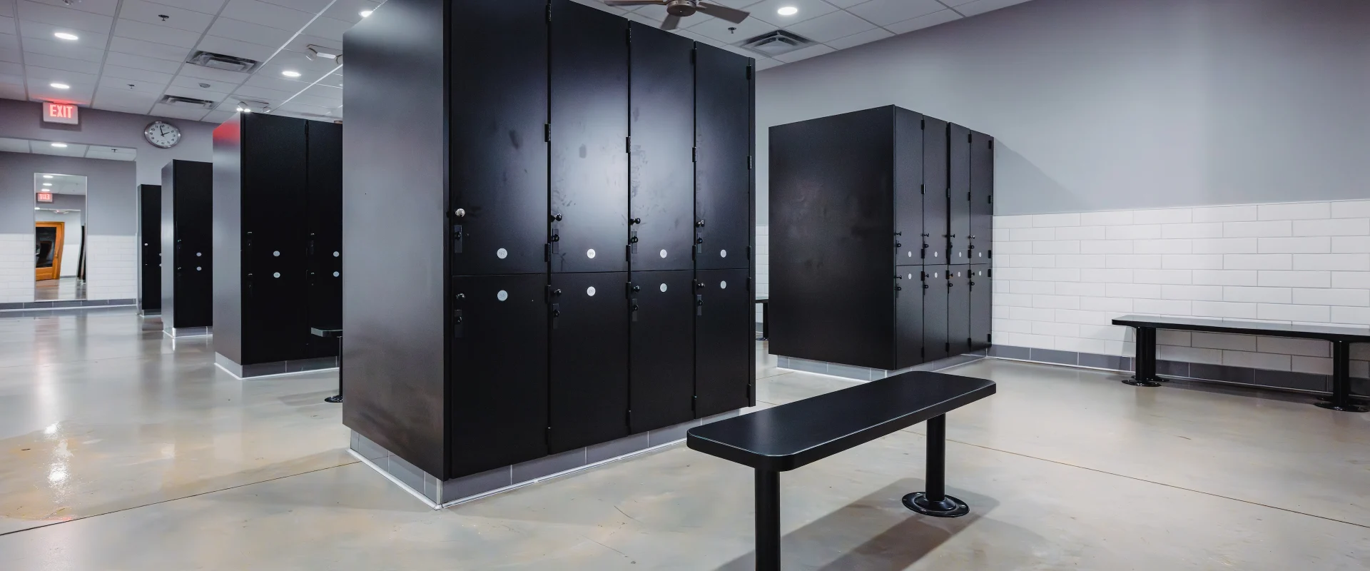 The locker room at Gold's Gym in Texas (Slaughter & South Congress) is equipped with black lockers, sturdy benches, and tiled floors. Overhead lighting ensures a bright, clean environment, while wall-mounted mirrors provide visibility in the grooming areas.