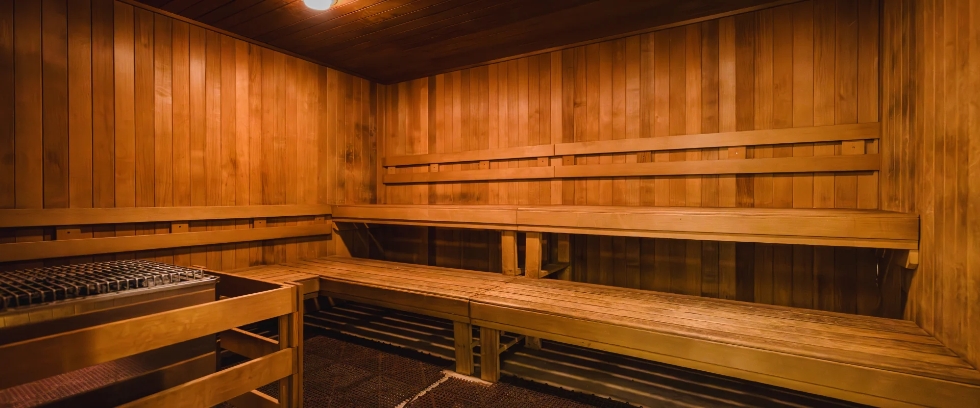 Dimly lit sauna room at Gold's Gym Austin (Anderson Arbor) with warm wood-paneled walls, matching wooden benches, and a dark, textured floor surrounding a corner heating element.