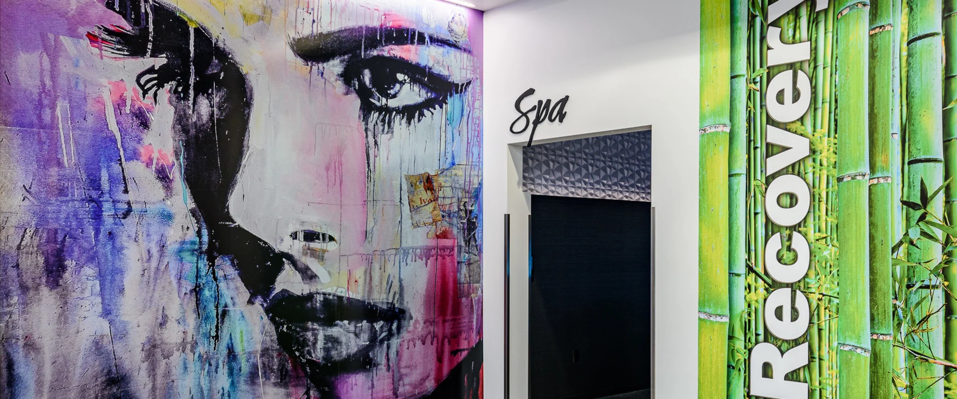 The recovery area at Gold’s Gym Cerritos features a spa entrance and a sign reading 'Spa' in cursive lettering. A large mural of a woman's face with abstract brushstrokes in purple, pink, and black covers one wall, while a vibrant green bamboo-themed panel with the word 'Recovery' in bold white letters decorates the adjacent wall. The polished concrete floor reflects soft overhead lighting.