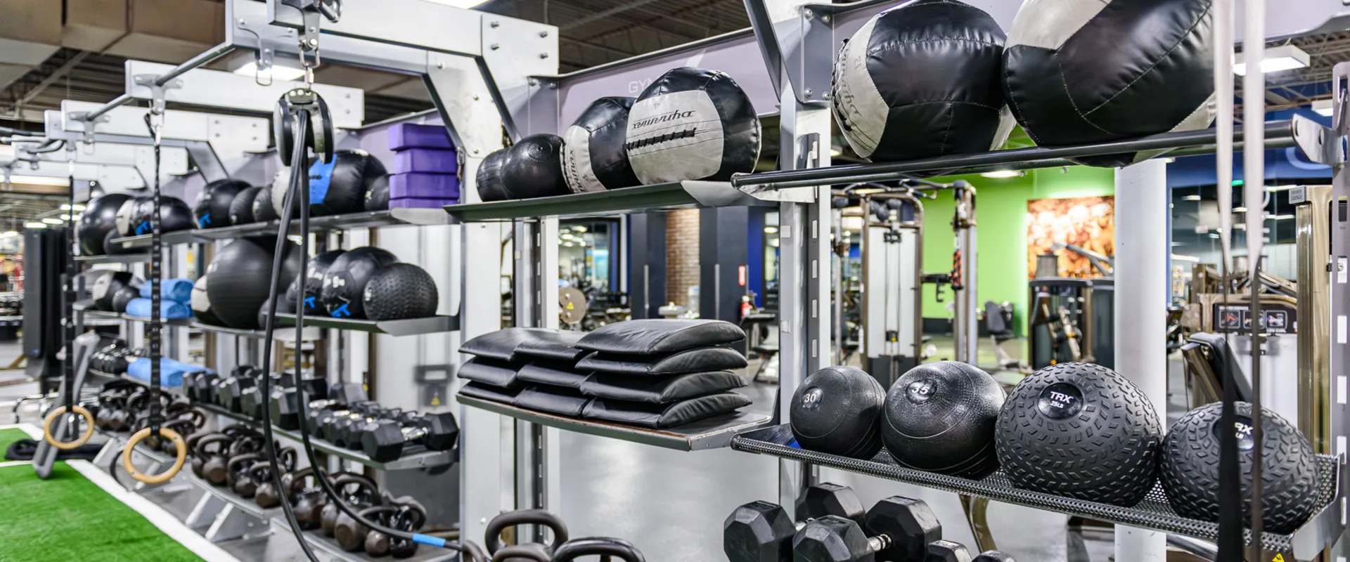 A multi-functional training station with shelves of medicine balls, kettlebells, resistance bands, and TRX straps sits adjacent to an artificial turf area with battle ropes and sled tracks.