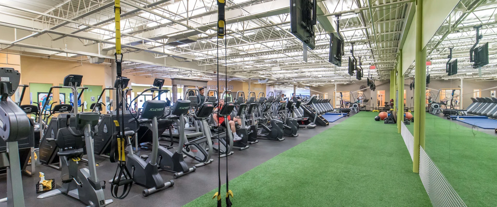 A large, well-equipped gym with various exercise equipment and machines arranged in rows on a green artificial turf floor, surrounded by high ceilings and exposed structural elements.