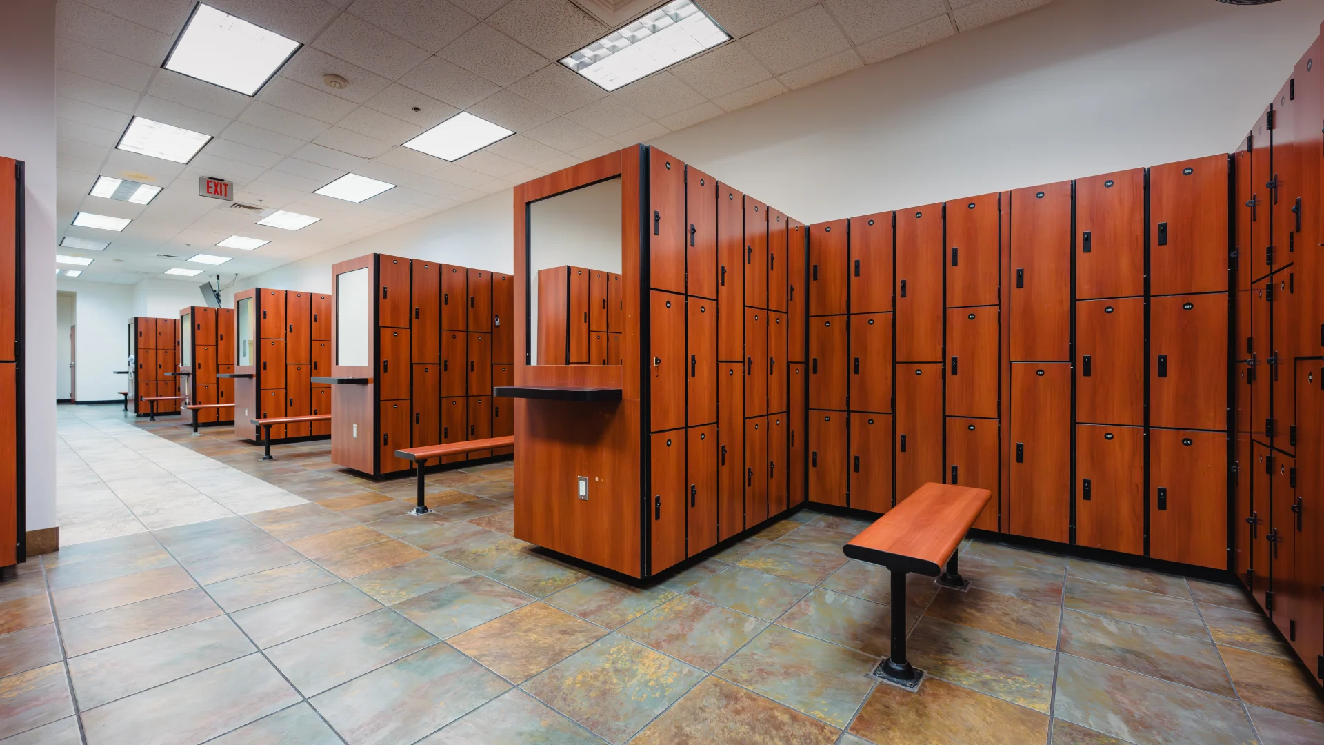 Locker room with rows of warm-toned wood lockers featuring black accents. Benches with matching wood tops and metal legs are positioned between the locker units. The room has a tiled floor in earthy tones, a drop ceiling with bright recessed lighting, and a clean, spacious layout. A wall-mounted fan is visible near the ceiling.