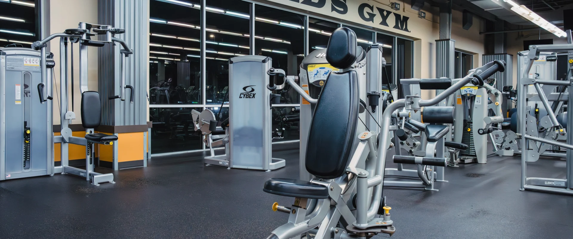 A modern gym interior with various exercise machines on a dark floor. Large "Gold's Gym" signage is visible above a mirrored wall, creating a clean, professional ambiance.
