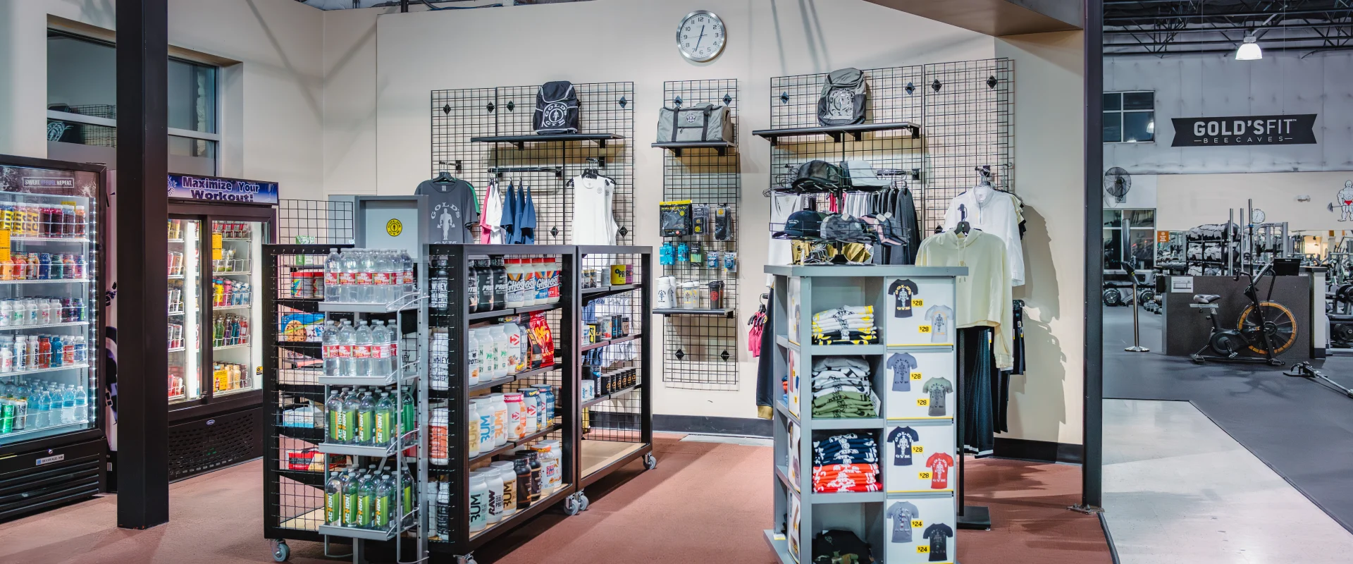 Gold's Gym pro shop in Austin (Bee Caves) showcasing refrigerators stocked with beverages and supplements, shelves filled with protein powders and energy drinks, and racks displaying branded apparel such as t-shirts, hats, and gym bags. The space is well-lit with a wall-mounted clock, adjacent to an open view of the gym floor labeled "Gold's Fit."