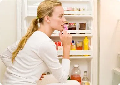 A young woman with long blonde hair stands in front of an open refrigerator, appearing to contemplate her food choices.