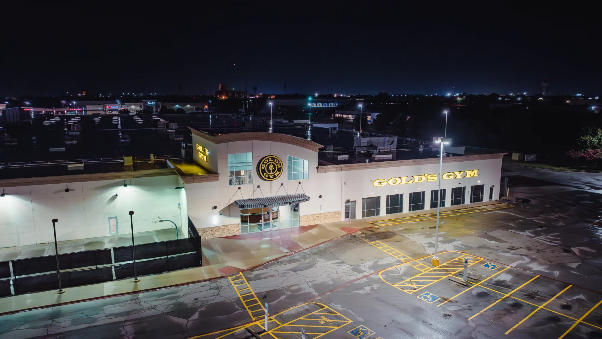 Nighttime exterior of Gold's Gym Waco featuring two illuminated logos, a well-lit entrance with modern architecture, empty parking spaces marked with accessible signage, and surrounding city lights in the background.