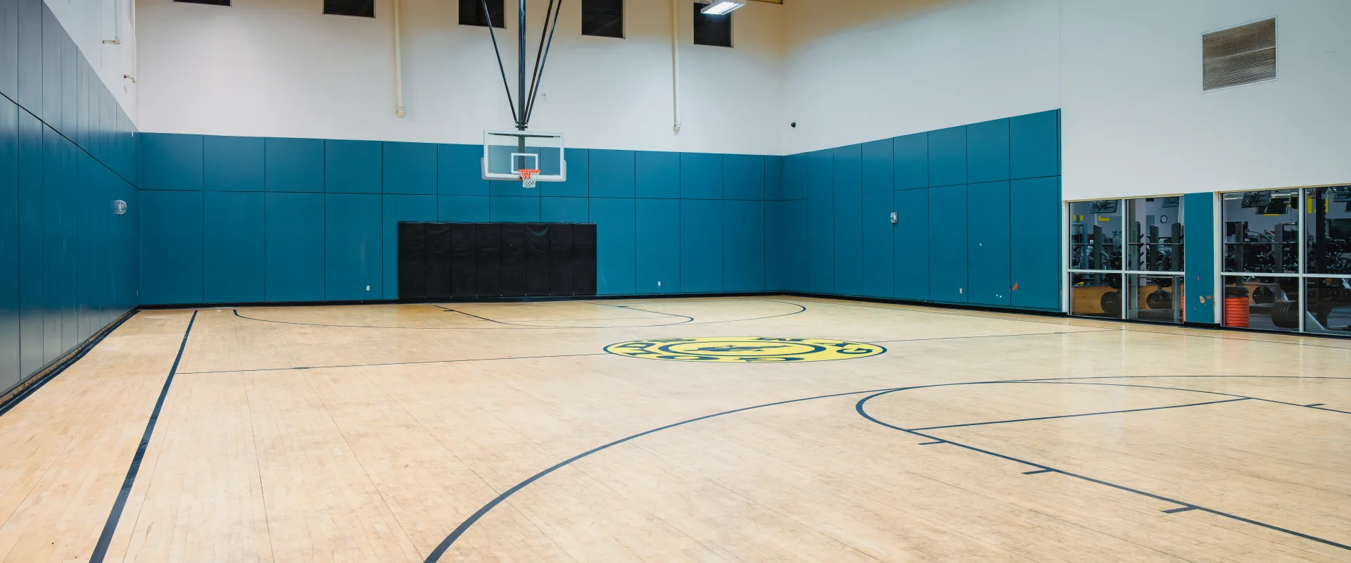 A basketball court at Gold's Gym in Austin (Hesters Crossing) featuring a polished wooden floor with court markings, including the gym's logo at the center. The walls are padded in blue with a mounted basketball hoop on one end, and high ceilings with bright overhead lights illuminate the space. A glass wall on the right side provides a view into the adjacent fitness area.