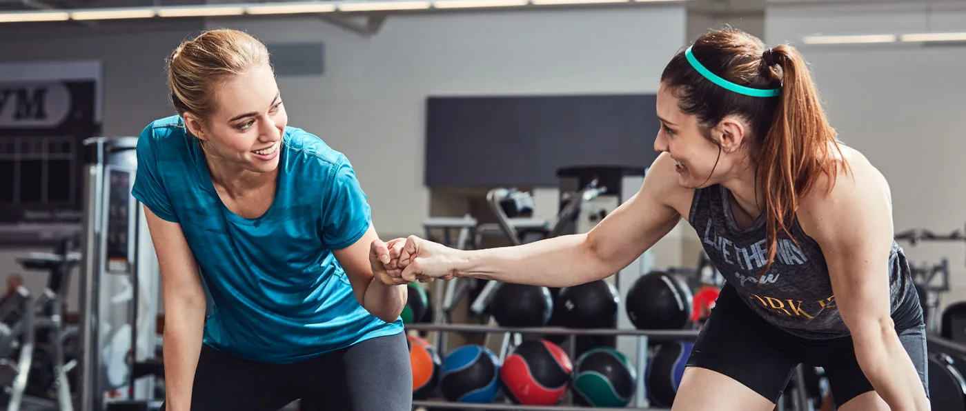 Two women in workout attire are interacting with each other in a gym setting, with various exercise equipment visible in the background.