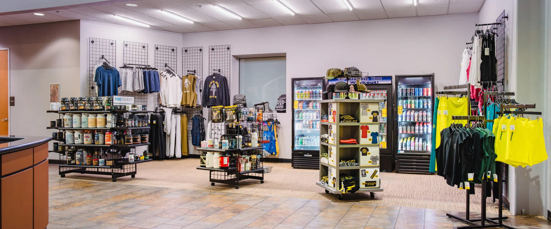 Pro shop at Gold's Gym Waco featuring a variety of fitness merchandise, including supplements, apparel, and accessories displayed on shelves and racks. Refrigerated coolers stock drinks and protein snacks, while tile flooring and bright lighting enhance the retail space.