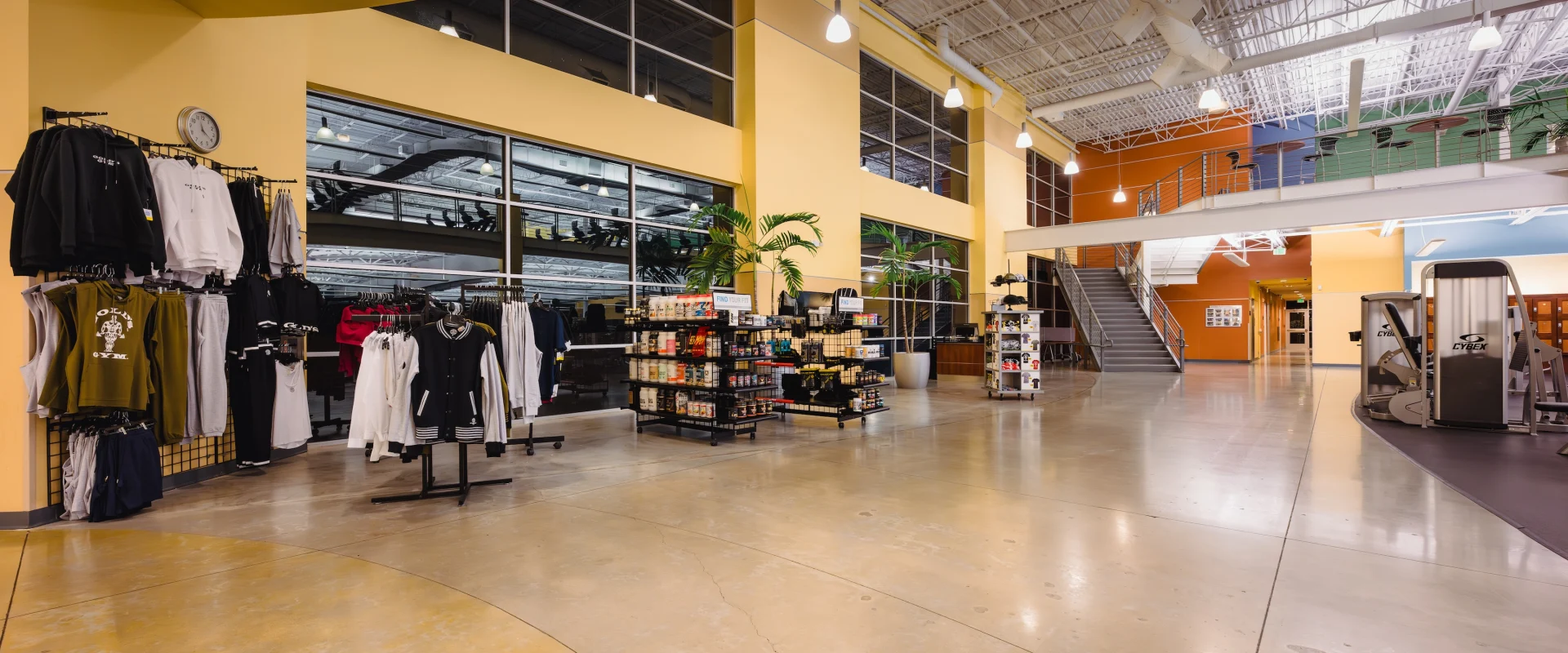 Wide gym lobby with yellow walls, clothing racks on the left, a staircase leading to the upper level, potted plants, and display shelves in the center. Bright lighting adds a welcoming atmosphere.