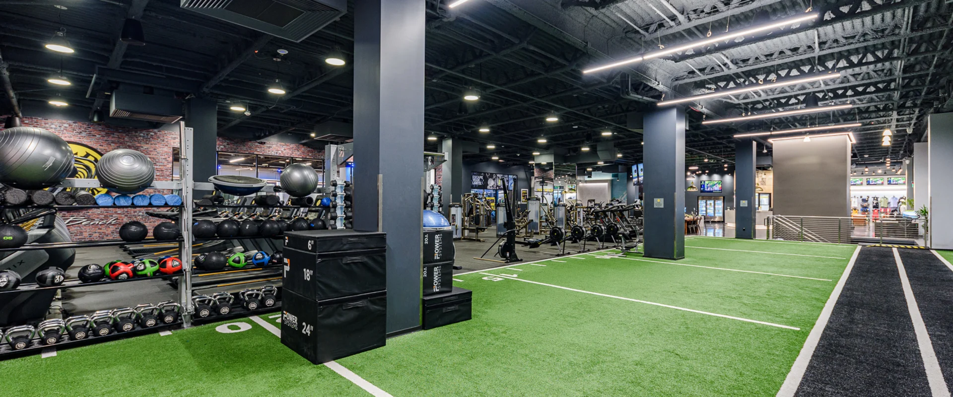 Turf area at Gold’s Gym Long Beach, designed for functional training and athletic conditioning. The space features green turf lanes marked for sled pushes and agility drills, alongside a black track for sprint work. A variety of equipment, including plyometric boxes, kettlebells, medicine balls, and battle ropes, is neatly organized on racks. Overhead industrial lighting and an open floor plan create a dynamic and spacious workout environment. In the background, cardio machines, weightlifting stations, and a pro shop are visible, offering a full-service gym experience.